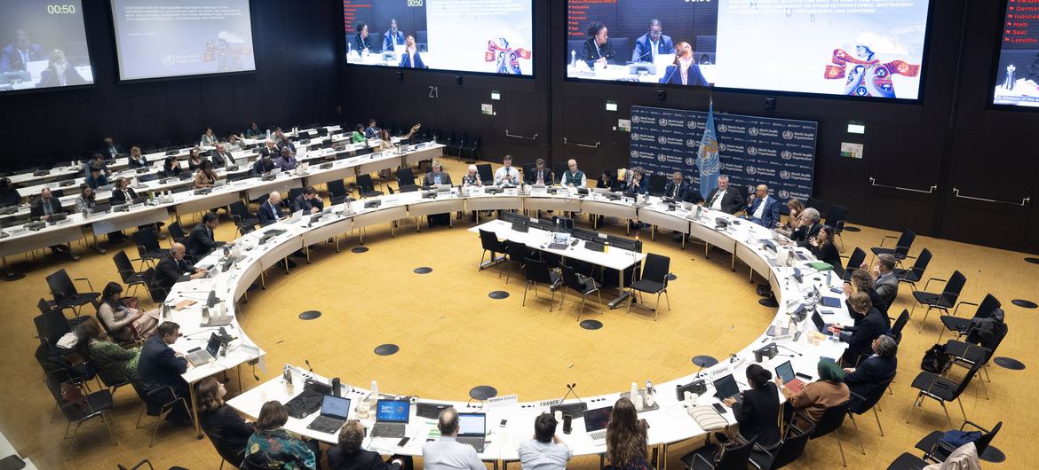 Les délégués sont assis autour d'une grande table de conférence en forme de U dans une salle de réunion formelle, avec plusieurs écrans vidéo au mur.